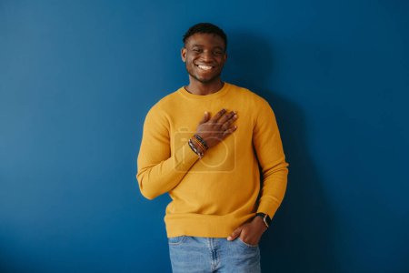 Téléchargez les photos : Heureux jeune homme africain tenant la main sur le cœur et regardant reconnaissant tout en se tenant sur fond bleu - en image libre de droit