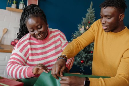Foto de Feliz joven pareja africana envolviendo regalos de Navidad en papel colorido en casa juntos - Imagen libre de derechos