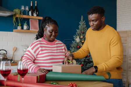 Photo for Beautiful young African couple wrapping Christmas gifts in colorful paper at home together - Royalty Free Image