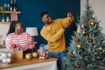 Foto de Hermosa pareja africana joven mirando feliz mientras decoran el árbol de Navidad en casa juntos - Imagen libre de derechos