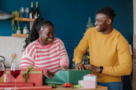 Foto de Hermosa pareja africana joven buscando alegre mientras envuelve los regalos de Navidad en casa juntos - Imagen libre de derechos