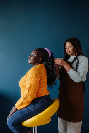 Téléchargez les photos : Coiffeur africain confiant tressant les cheveux à une cliente sur fond bleu - en image libre de droit