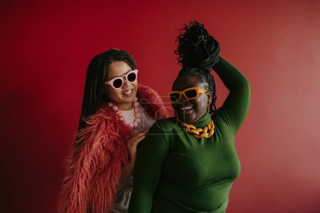 Téléchargez les photos : Deux joyeux plus la taille femme africaine jouer avec leurs cheveux tressés tout en s'amusant sur fond rouge - en image libre de droit