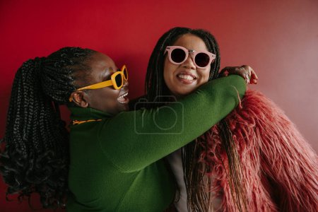 Foto de Dos juguetones más tamaño mujer africana con pelo trenzado abrazando sobre fondo rojo juntos - Imagen libre de derechos