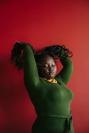 Téléchargez les photos : Belle femme africaine incurvée ajustant ses cheveux tressés et gardant les yeux fermés sur fond rouge - en image libre de droit
