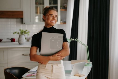 Téléchargez les photos : Attrayant jeune femme tenant ordinateur portable et souriant tout en se tenant debout au bureau à la maison - en image libre de droit