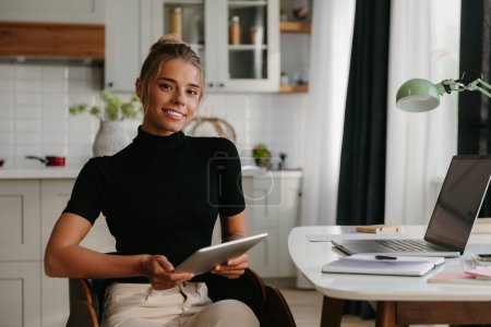 Téléchargez les photos : Une jeune femme confiante tenant une tablette numérique et souriant à la caméra alors qu'elle était assise au bureau à la maison - en image libre de droit