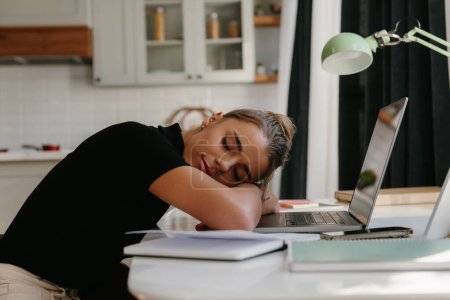 Foto de Joven mujer de negocios cansada apoyada de cabeza en el portátil y durmiendo mientras está sentada en el escritorio en casa - Imagen libre de derechos