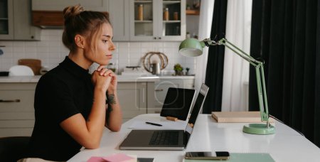 Téléchargez les photos : Une jeune femme concentrée assise au bureau et regardant un ordinateur portable tout en travaillant à la maison - en image libre de droit