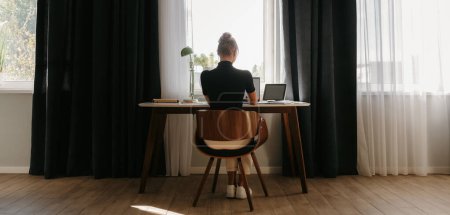 Photo for Rear view of young businesswoman working at home while sitting at the desk in front of the window - Royalty Free Image