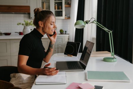 Téléchargez les photos : Une jeune femme confiante parle sur un téléphone portable et regarde un ordinateur portable tout en travaillant à la maison - en image libre de droit