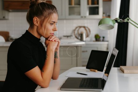 Téléchargez les photos : Jeune femme réfléchie assise au bureau et regardant un ordinateur portable tout en travaillant à la maison - en image libre de droit
