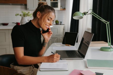 Foto de Mujer joven y feliz hablando por teléfono móvil y tomando notas mientras trabaja en casa - Imagen libre de derechos
