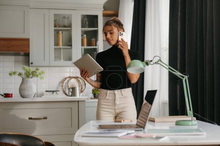 Téléchargez les photos : Concentré jeune femme parlant sur téléphone portable et en utilisant une tablette numérique tout en travaillant à la maison - en image libre de droit
