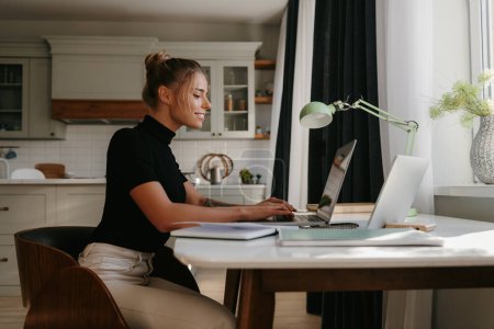 Téléchargez les photos : Vue latérale d'une jolie jeune femme assise au bureau et utilisant un ordinateur portable tout en travaillant à la maison - en image libre de droit
