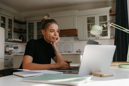 Foto de Mujer joven aburrida sentada en el escritorio y mirando a la computadora portátil mientras estudia en casa - Imagen libre de derechos
