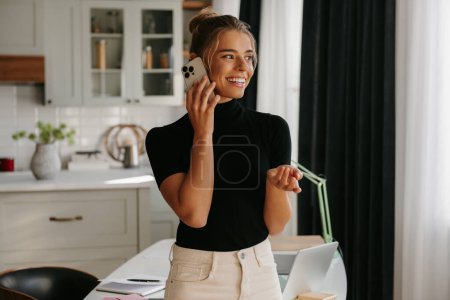 Téléchargez les photos : Une jeune femme confiante parlant sur son téléphone portable et souriant debout au bureau à la maison - en image libre de droit