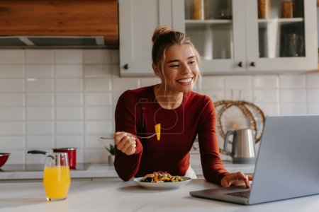 Foto de Hermosa mujer joven disfrutando de un almuerzo saludable y trabajando en el ordenador portátil en la cocina doméstica - Imagen libre de derechos
