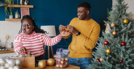 Photo for Beautiful young African couple looking happy while decorating Christmas tree at home together - Royalty Free Image