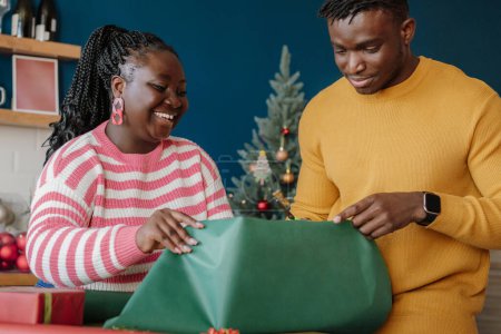 Foto de Joven pareja africana alegre envolviendo regalos de Navidad en papel colorido en casa juntos - Imagen libre de derechos