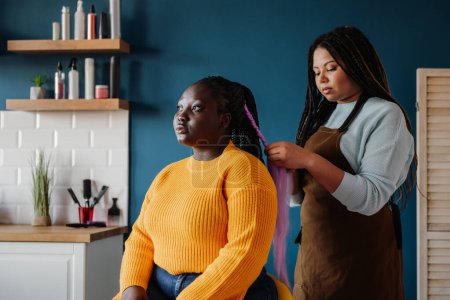 Foto de Peluquería afroamericana confiada trenzando el cabello a una cliente femenina en el salón - Imagen libre de derechos