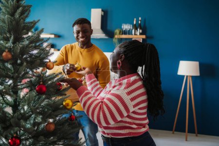 Photo for Joyful young African couple enjoying fun time while decorating Christmas tree at home together - Royalty Free Image