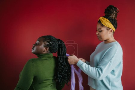 Téléchargez les photos : Coiffeur africain confiant tressant les cheveux à une belle jeune femme sur fond rouge - en image libre de droit