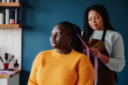 Téléchargez les photos : Coiffeur africain confiant tresser les cheveux à la cliente dans le salon - en image libre de droit