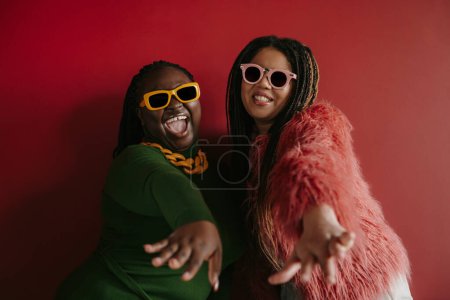 Téléchargez les photos : Deux joyeux plus la taille femme africaine avec les cheveux tressés étirant les mains sur fond rouge ensemble - en image libre de droit