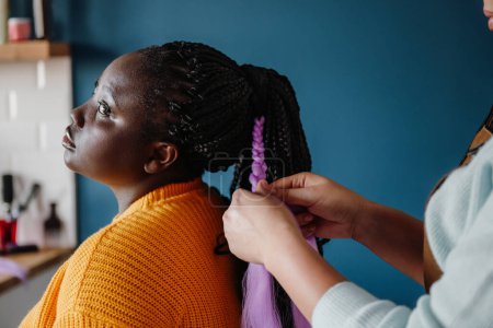 Foto de Primer plano de peluquería trenzando el pelo a una joven mujer africana en el salón - Imagen libre de derechos