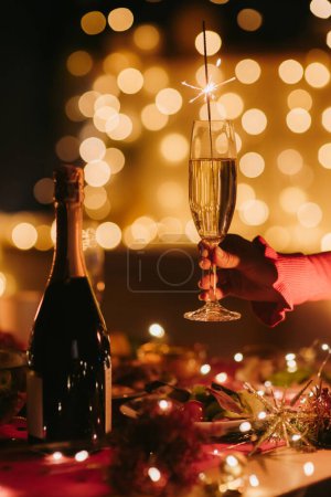 Foto de La mano femenina sostiene la flauta con champán mientras celebra el Año Nuevo en la mesa de fiesta decorada - Imagen libre de derechos