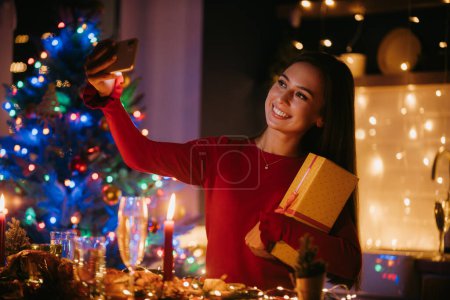 Foto de Mujer joven sonriente sosteniendo caja de regalo y haciendo selfie mientras disfruta de la víspera de Año Nuevo en casa - Imagen libre de derechos