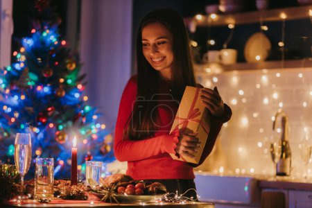Foto de Mujer joven sonriente sosteniendo caja de regalo mientras disfruta de la víspera de Año Nuevo en la habitación decorada en casa - Imagen libre de derechos
