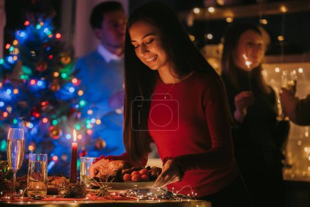 Foto de Mujer joven sonriente sirviendo platos navideños en la mesa mientras sus amigos celebran el Año Nuevo en segundo plano - Imagen libre de derechos