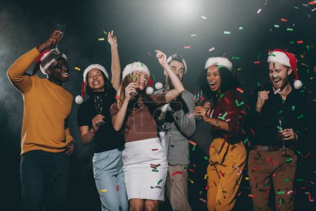 Téléchargez les photos : Des jeunes gens joyeux dans des chapeaux de Noël dansant et jetant des confettis tout en célébrant le Nouvel An en boîte de nuit - en image libre de droit