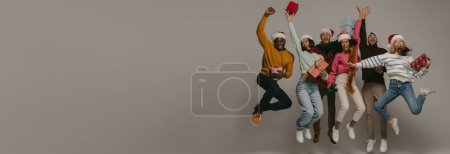 Photo for Happy young people carrying Christmas ornaments and gift boxes while jumping on background together - Royalty Free Image
