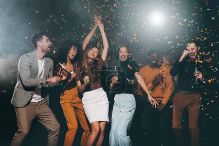 Photo for Group of young happy people enjoying champagne and throwing confetti while dancing in night club together - Royalty Free Image