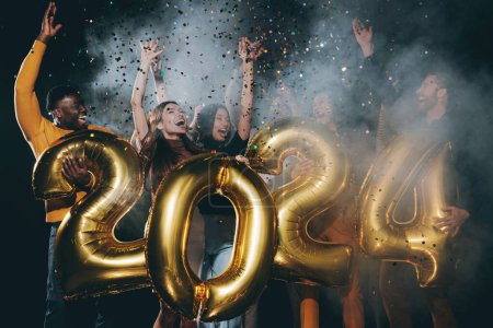 Photo for Group of joyful people holding 2024 numbers and throwing confetti while celebrating New Year in night club - Royalty Free Image