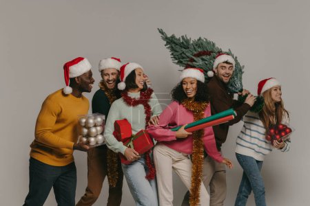 Foto de Jóvenes felices llevando el árbol de Navidad y varios ornamentos mientras caminan juntos sobre el fondo del estudio - Imagen libre de derechos