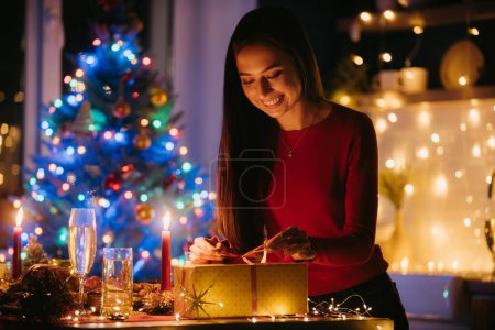 Téléchargez les photos : Souriant jeune femme ouverture boîte cadeau tout en profitant du Nouvel An dans la chambre décorée à la maison - en image libre de droit