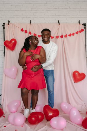 Téléchargez les photos : Pleine longueur de heureux jeune couple africain embrassant sur fond rose avec des ballons en forme de coeur - en image libre de droit
