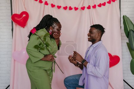 Foto de Hombre africano confiado dando anillo de compromiso a su novia sorprendida mientras hace propuesta sobre fondo rosa - Imagen libre de derechos