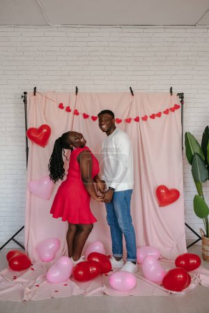 Foto de Sonriente joven pareja africana cogida de la mano mientras está de pie sobre fondo de tela rosa con globos en forma de corazón - Imagen libre de derechos