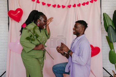 Téléchargez les photos : Jeune homme africain donnant bague de fiançailles à sa petite amie surprise tout en faisant une proposition sur fond rose - en image libre de droit