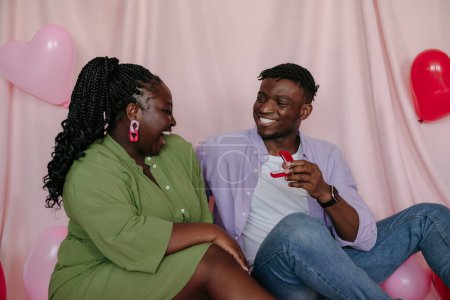 Foto de Feliz joven africano dando anillo de compromiso a su novia y pidiendo casarse con él en el fondo de tela rosa - Imagen libre de derechos