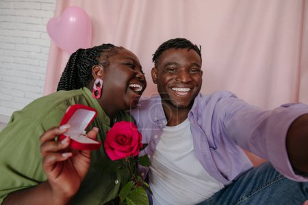 Téléchargez les photos : Joyeuse jeune femme africaine tenant la bague de fiançailles tout en embrassant son petit ami sur fond rose - en image libre de droit