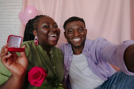 Téléchargez les photos : Heureuse jeune femme africaine tenant la bague de fiançailles tout en embrassant son petit ami sur fond rose - en image libre de droit
