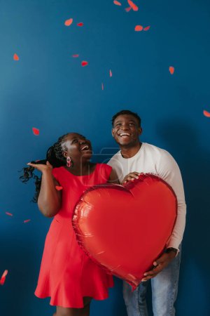 Foto de Feliz joven pareja africana sosteniendo globo en forma de corazón rojo mientras confeti volando sobre fondo azul - Imagen libre de derechos
