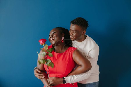 Foto de Feliz joven africano abrazando a su novia sosteniendo rosa roja sobre fondo azul - Imagen libre de derechos