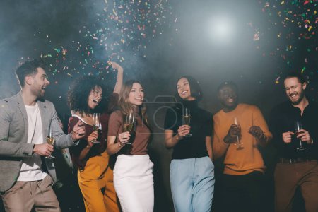 Téléchargez les photos : Groupe de jeunes souriants profitant de la fête dans la boîte de nuit avec des confettis volant autour - en image libre de droit